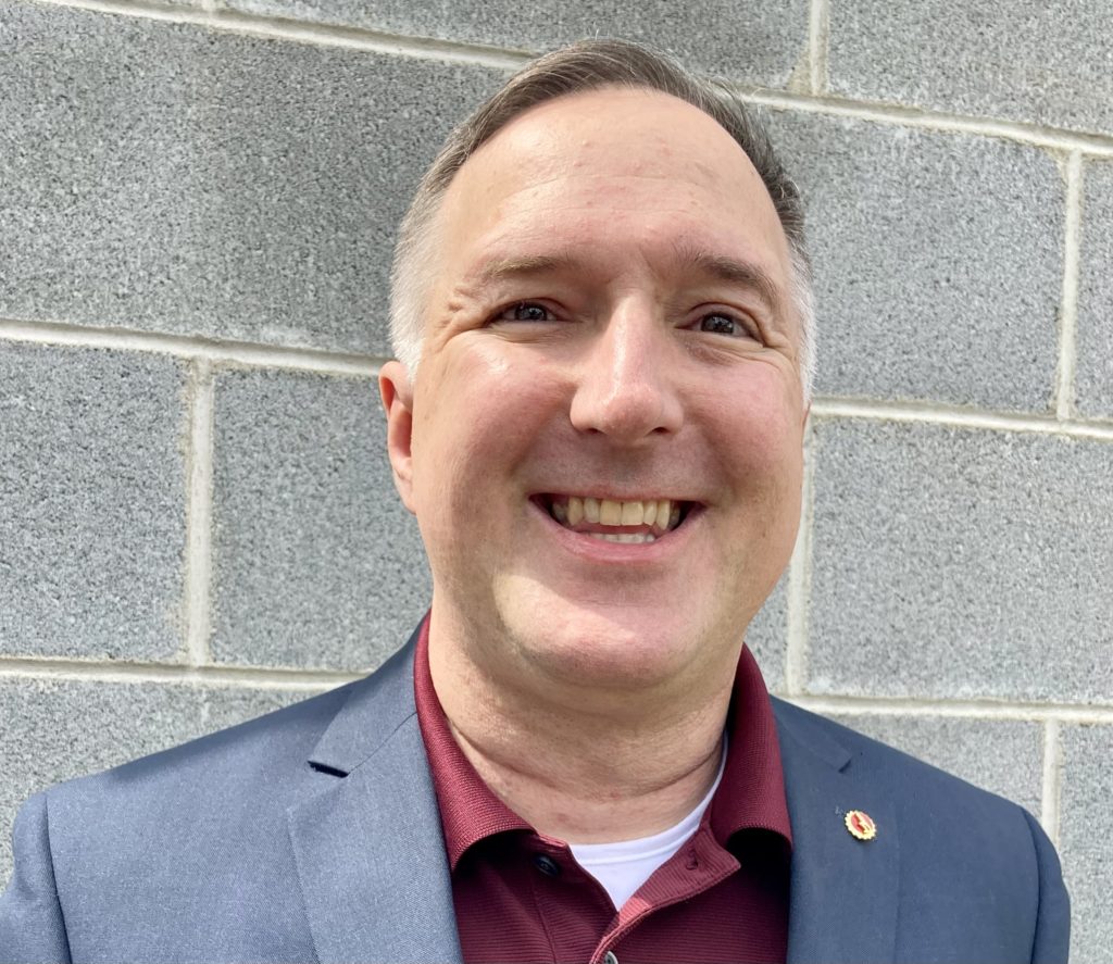 Jeff Price standing against a stone backdrop smiling with a blue blazer, minute man pin, red polo,shirt, t-white shirt 